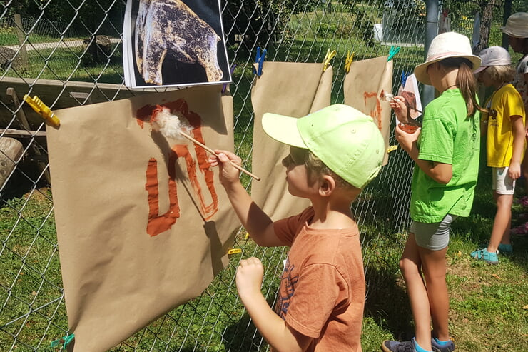 Mehrere Kinder malen im Freien mit Pinseln auf braunes Papier, das an einem Zaun befestigt ist.