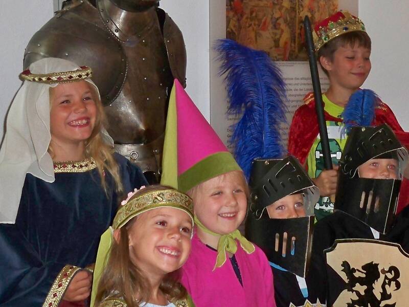 Eine Gruppe von Kindern in historischen Kostümen steht lächelnd vor einer Ritterrüstung.