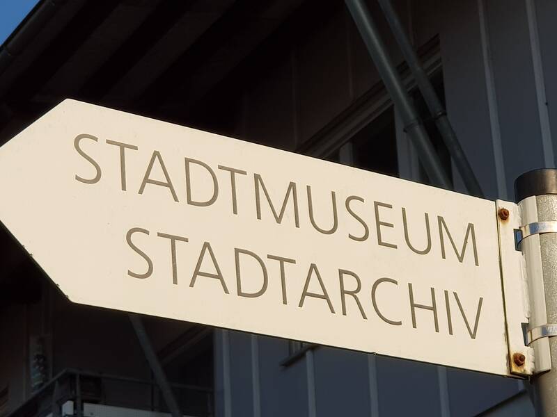 Ein weißes Schild auf einem Metallpfosten zeigt in Richtung Stadtmuseum und Stadtarchiv.