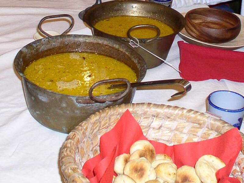 Zwei Töpfe mit gelber Suppe stehen auf einem Tisch neben einem geflochtenen Korb mit kleinen Brötchen auf einem roten Tuch.