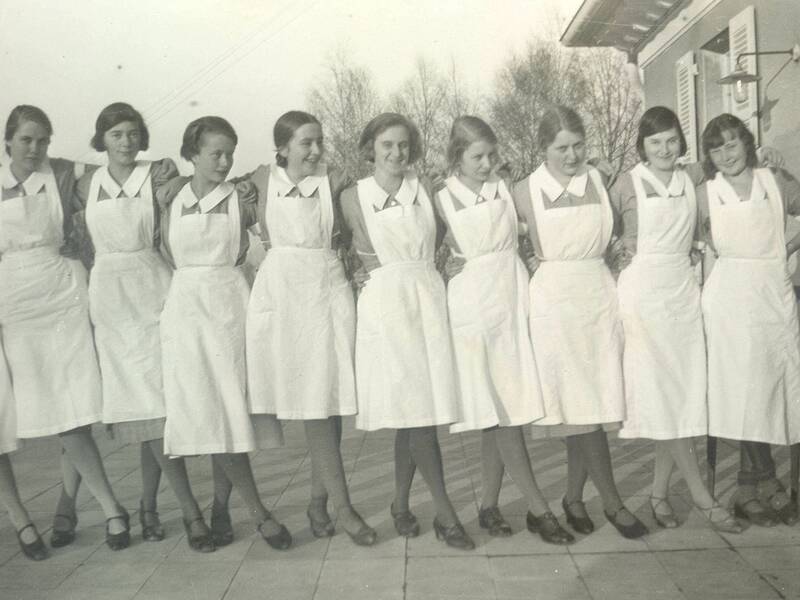Maiden 1931 / Eine Gruppe von zehn Frauen in weißen Schürzen steht dicht nebeneinander aufgereiht und lächelt in die Kamera.