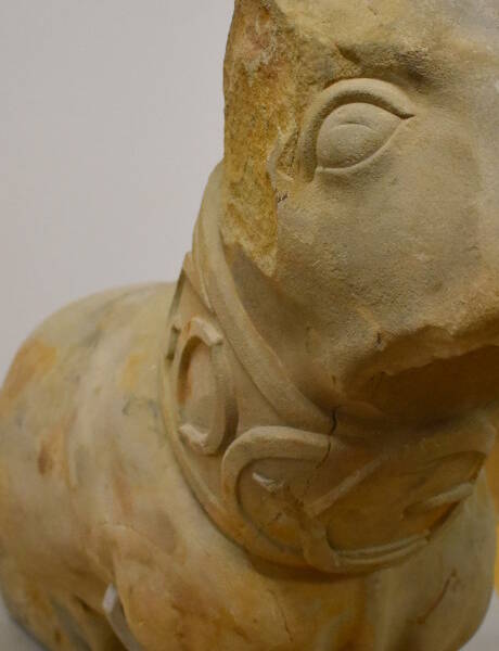 Steinhund / Eine grob behauene Steinskulptur zeigt einen stilisierten Tierkopf mit einem dekorativen Halsband und glatten, abgerundeten Formen.