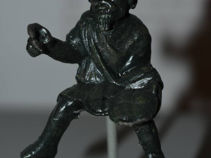Eine römische Bronzestatuette / Eine kleine, dunkle Statue eines sitzenden Mannes mit Bart und lockigem Haar, der einen Gegenstand in der rechten Hand hält.