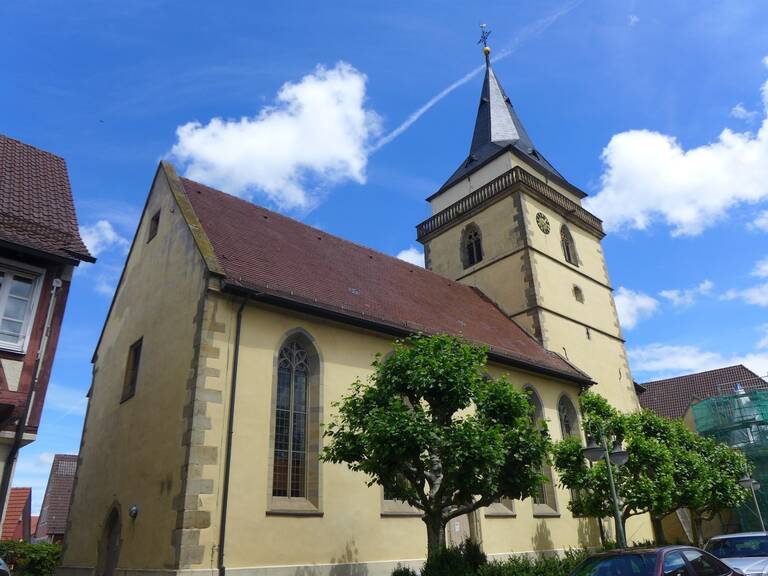 Das Bild zeigt eine Kirche mit einem spitzen Turm und Uhr, umgeben von Bäumen und einem klaren blauen Himmel.