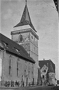 Das Bild zeigt eine historische Kirche mit einem hohen Kirchturm und mehreren Personen, die vor dem Gebäude stehen.