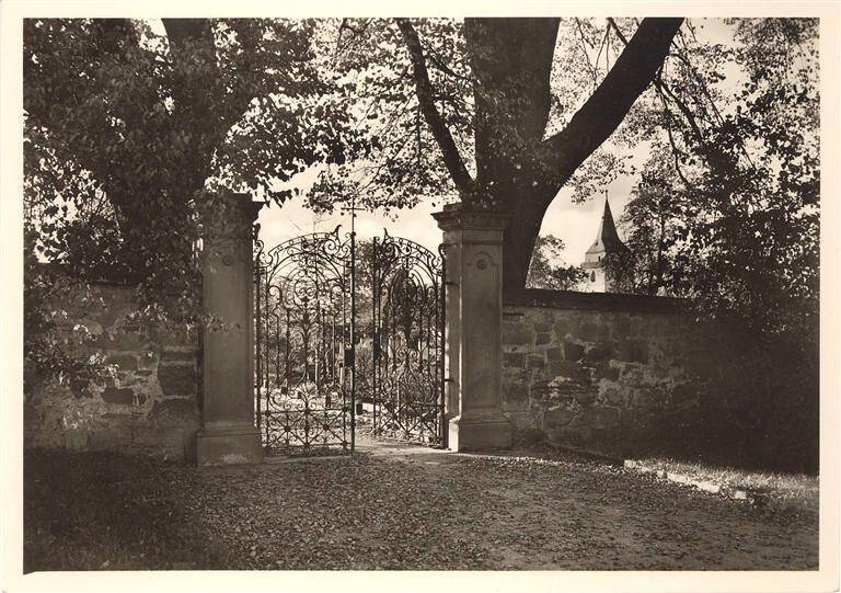 Eine kunstvoll geschmiedete Toranlage steht zwischen alten Bäumen und einer Steinmauer, mit einem Kirchturm im Hintergrund.