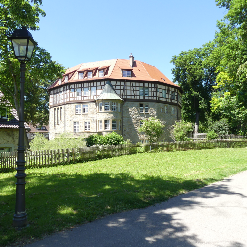 Das Wasserschloss mit rotem Dach steht in einem grünen, parkähnlichen Umfeld mit Laternen und Bäumen.