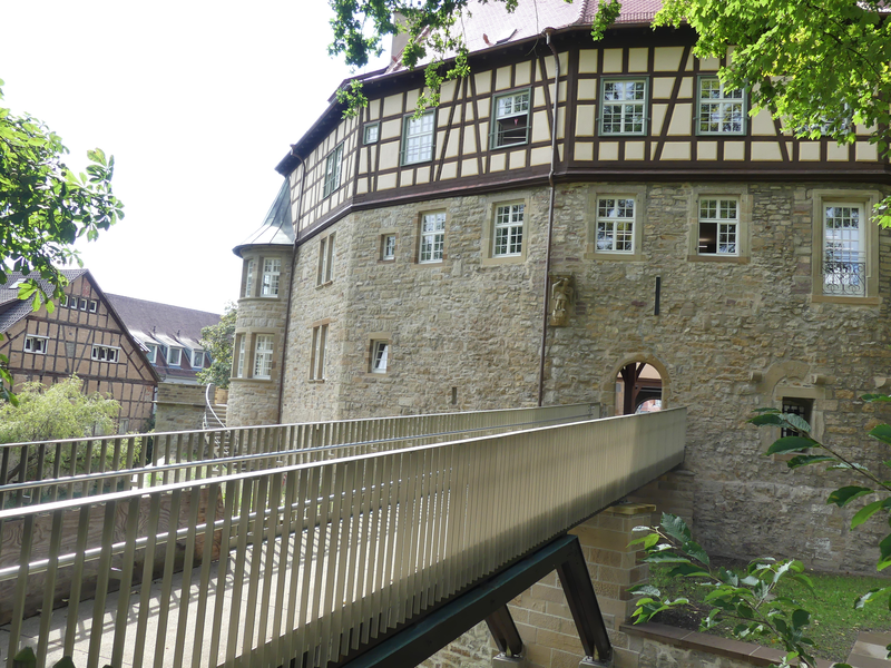Ein historisches Fachwerkgebäude mit einer Steinmauer ist durch eine moderne Brücke erreichbar, umgeben von Bäumen.