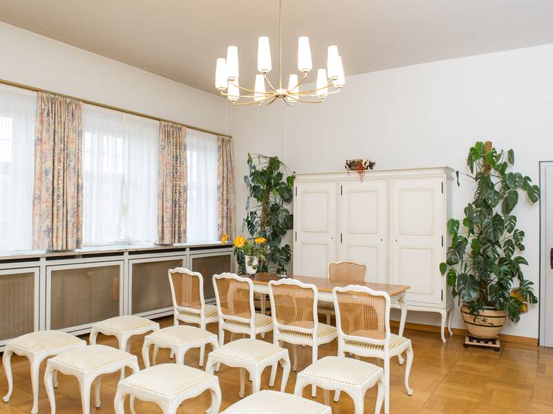 Ein heller Raum mit weißen Möbeln, darunter ein Tisch mit Stühlen und Hockern, einem großen Schrank sowie Zimmerpflanzen und einem Leuchter an der Decke.