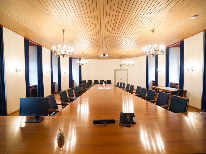 Ein großer Konferenzraum mit einer langen Holztafel, umgeben von vielen Stühlen und beleuchtet durch mehrere Kronleuchter.