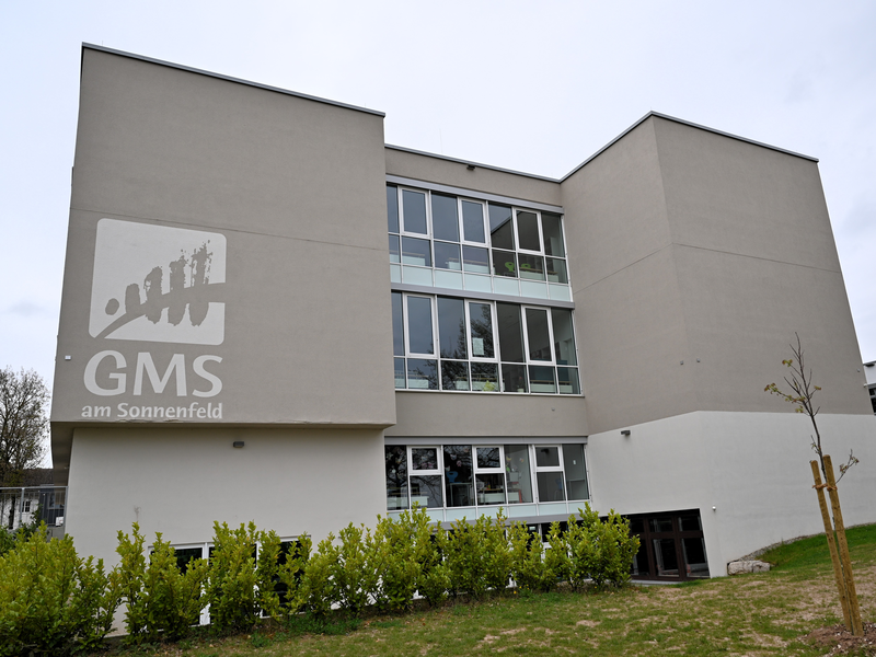 Ein modernes, mehrstöckiges Gebäude mit großen Fenstern und einem Logo der "GMS am Sonnenfeld" an der Fassade, umgeben von grünem Rasen und Sträuchern.