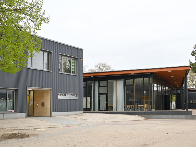 Das Bild zeigt ein modernes Gebäude mit großen Glasfenstern und einem flachen Dach, vor dem sich ein gepflasterter Vorplatz erstreckt, umgeben von Bäumen.