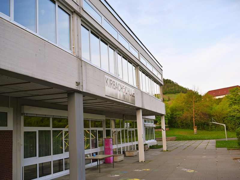 Das Bild zeigt die Außenansicht eines Schulgebäudes mit großen Fenstern und einem Eingang, umgeben von grüner Vegetation.