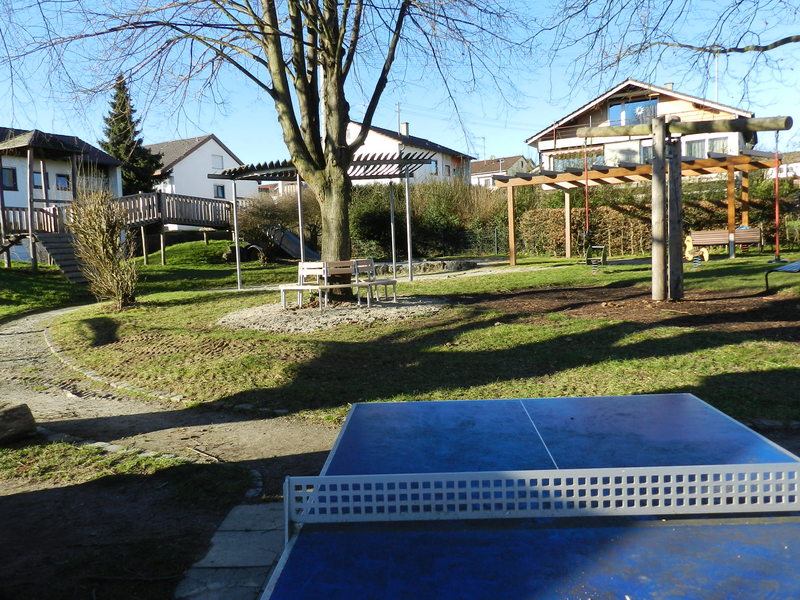 Ein Spielplatz in einer Wohngegend mit Klettergerüst, Bänken, einem Tischtennistisch und einem großen Baum im Hintergrund.