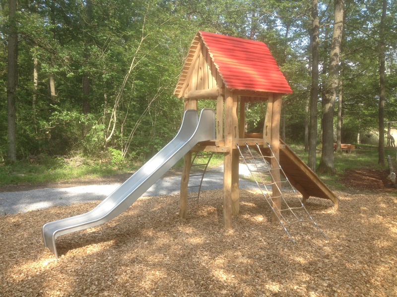 Ein Holzspielplatz mit einem roten Dach, Rutsche und Klettergerüst steht in einem bewaldeten Bereich.