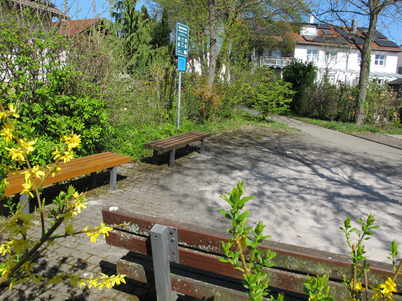 Das Bild zeigt einen sonnigen Park mit Bänken, einem Weg, dichten Büschen und blühenden gelben Blumen im Vordergrund.