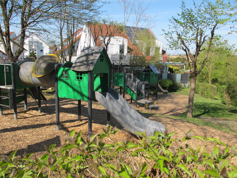 Ein Spielplatz mit grünen Spielhäusern, Rutschen und Klettergerüsten steht unter Bäumen in einer sonnigen Umgebung mit Häusern im Hintergrund.