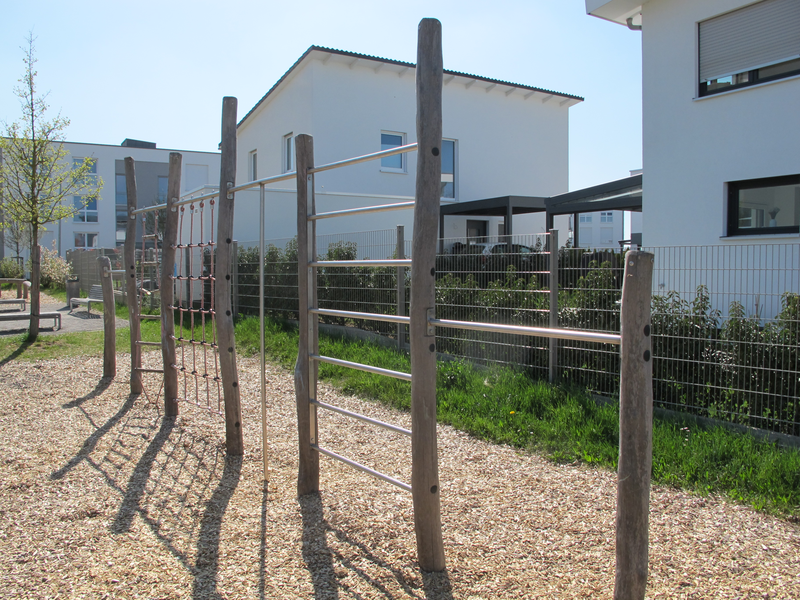 Ein Klettergerüst aus Seilen und Holzstämmen steht auf einem mit Holzschnitzeln bedeckten Spielplatz vor modernen weißen Häusern.