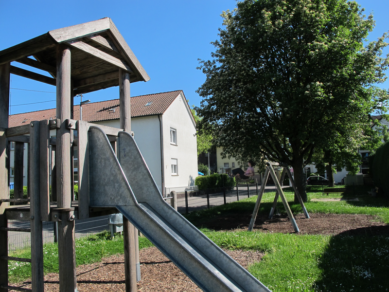 Ein Spielplatz mit einer Rutsche und einer Schaukel ist von Grünflächen und einem großen Baum umgeben, dahinter stehen Häuser unter einem klaren blauen Himmel.
