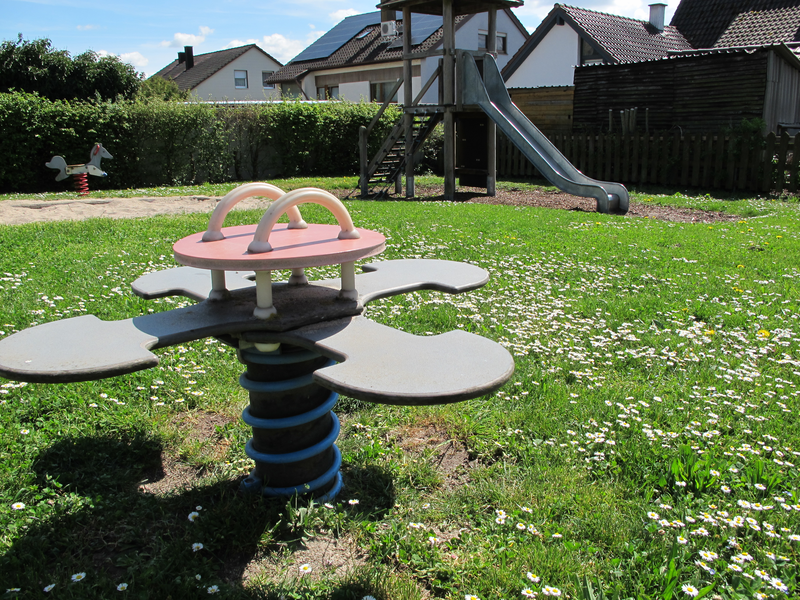 Ein Spielplatz mit einer Wippe im Vordergrund und einer Rutsche im Hintergrund, umgeben von einer grünen Wiese mit kleinen weißen Blumen.