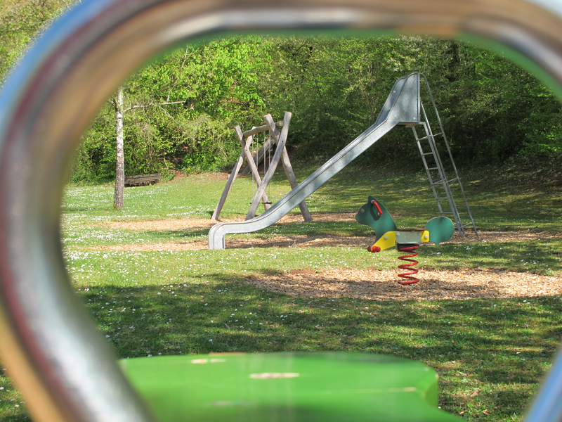 Ein Spielplatz mit einer Rutsche und einem wippenden Spielgerät in Form eines Tieres, gesehen durch einen Reifen.