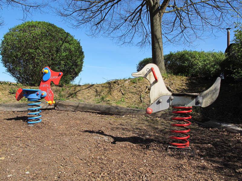 Auf einem Spielplatz stehen zwei schaukelnde Federwippen in Form eines Pferdes und einer Ente, umgeben von Bäumen und Sträuchern.
