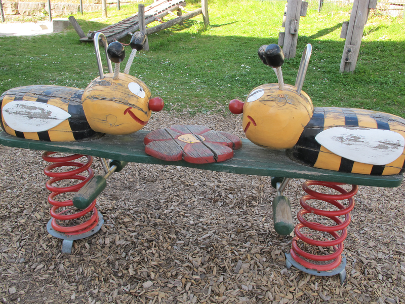Ein Wipptier in Form zweier gelber, schwarz gestreifter Bienen mit großen roten Nasen steht auf Spiralfedern auf einem Spielplatz.