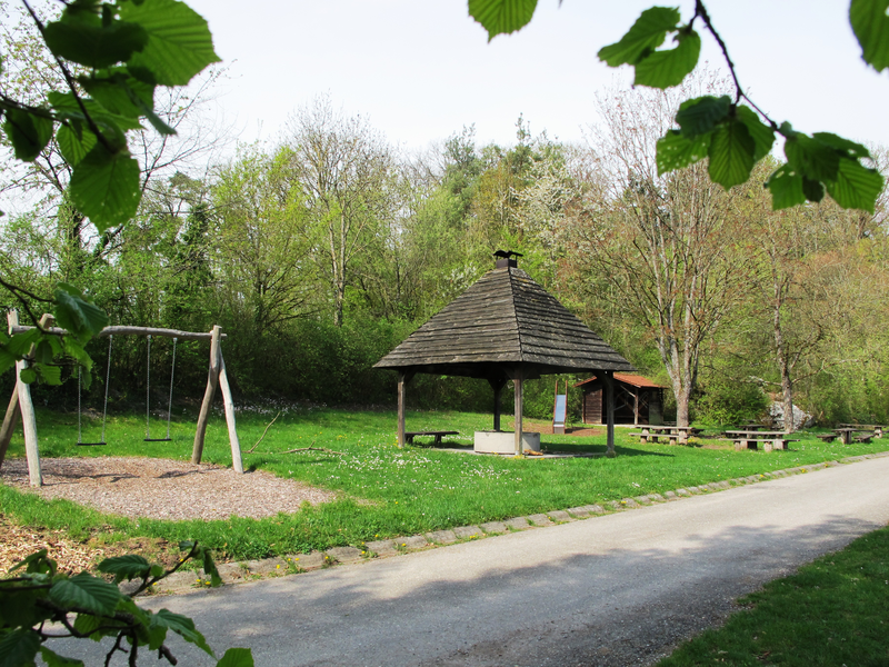 Ein Spielplatz mit Schaukel und ein überdachter Sitzbereich befinden sich neben einem Weg in einer grünen, bewaldeten Umgebung.