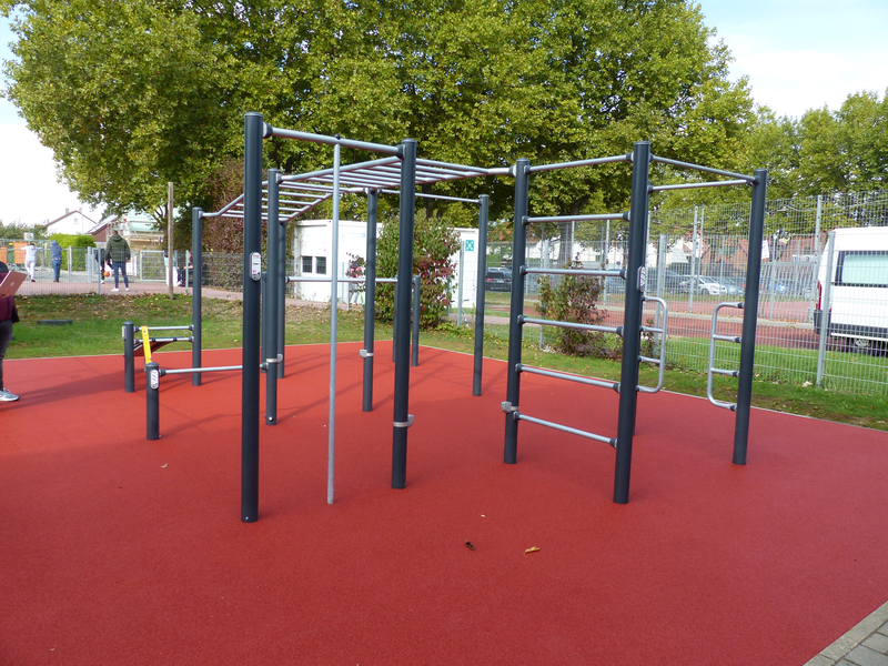 Ein moderner Outdoor-Spielplatz mit Klettergerüst auf rotem Boden ist von Bäumen und einem Zaun umgeben.