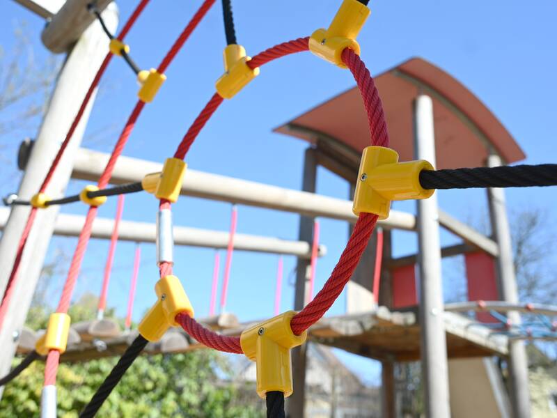 Ein Spielplatz-Klettergerüst mit roten Seilen und gelben Verbindungselementen vor einem klaren blauen Himmel.