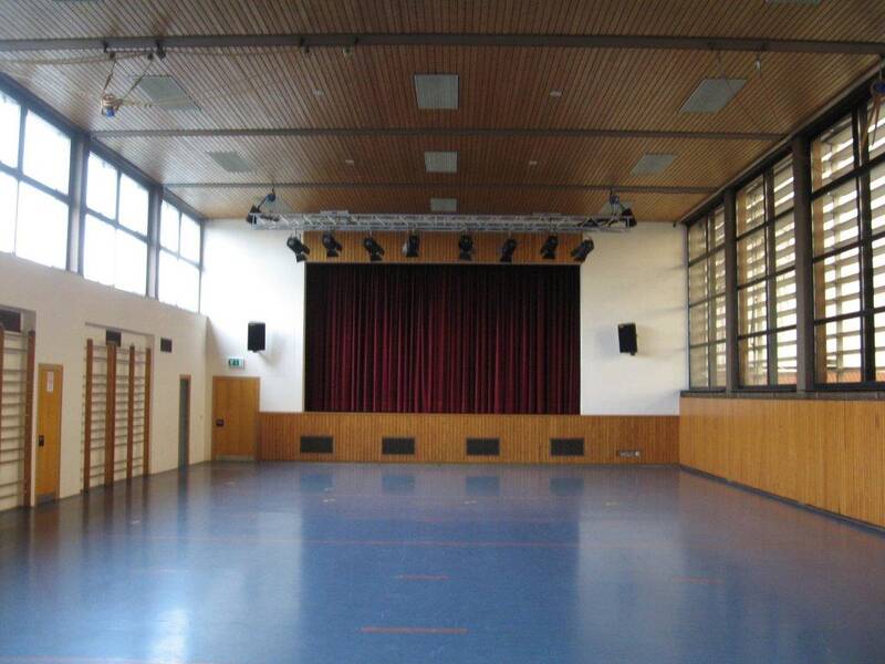 Ein leerer, heller Mehrzwecksaal mit großen Fenstern und einer Bühne mit einem roten Vorhang.