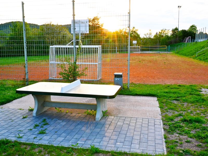 Eine Tischtennisplatte steht im Vordergrund auf einem gepflasterten Bereich vor einem umzäunten Sportplatz mit Basketballkorb und Volleyballnetz im Hintergrund bei Sonnenuntergang.