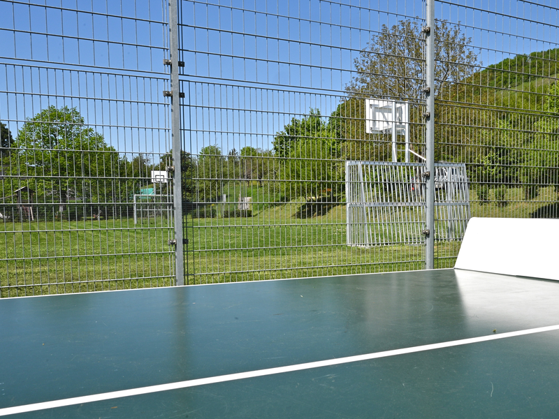 Im Vordergrund befindet sich eine Tischtennisplatte auf einem eingezäunten Sportplatz, im Hintergrund sind Basketballkörbe und grüne Bäume zu erkennen.
