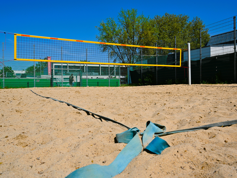 Ein Beachvolleyballfeld mit einem gelben Netz, umgeben von einem Zaun und im Hintergrund Bäumen, unter einem klaren blauen Himmel.