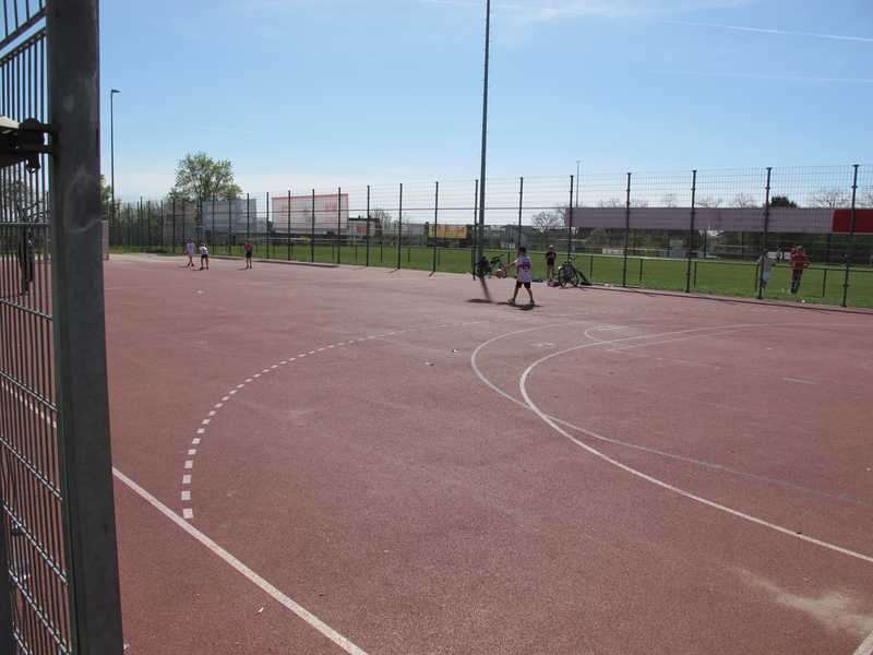 Kinder spielen auf einem umzäunten roten Sportplatz unter blauem Himmel.