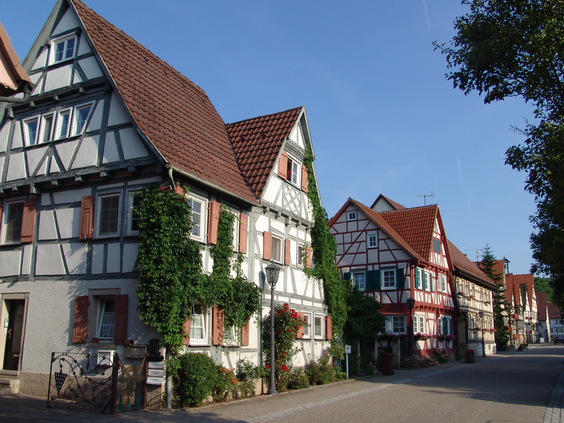 Eine Reihe traditioneller Fachwerkhäuser mit rotbraunen Dächern steht an einer ruhigen, von Bäumen gesäumten Straße.