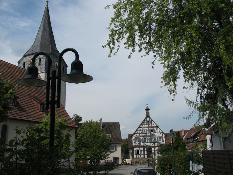 Eine malerische Dorfansicht zeigt eine Fachwerkhausfassade im Hintergrund, einen Kirchturm links und Straßenlaternen im Vordergrund, umgeben von Bäumen.