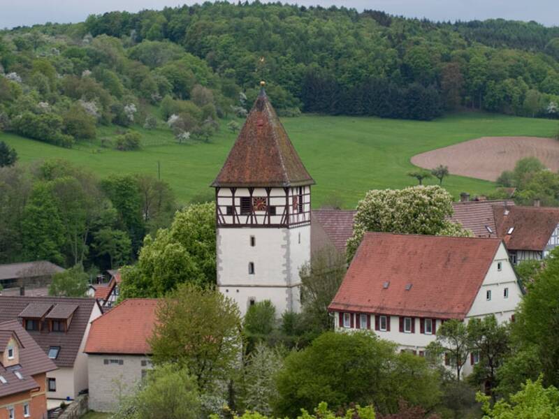 Ein malerisches Dorf mit einem historischen Turm, umgeben von üppigen grünen Hügeln und Wäldern.