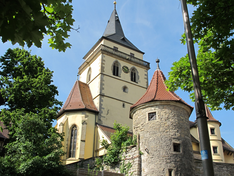 Eine historische Kirche mit einem hohen Turm und einer Rundbastion ist umgeben von grünen Bäumen, während ein blauer PKW im Vordergrund am Straßenrand parkt.