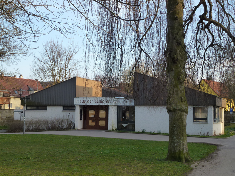 Ein flacher, moderner Gebäudekomplex mit der Aufschrift "Haus der Senioren" steht vor einem Rasen mit einem großen Baum im Vordergrund.