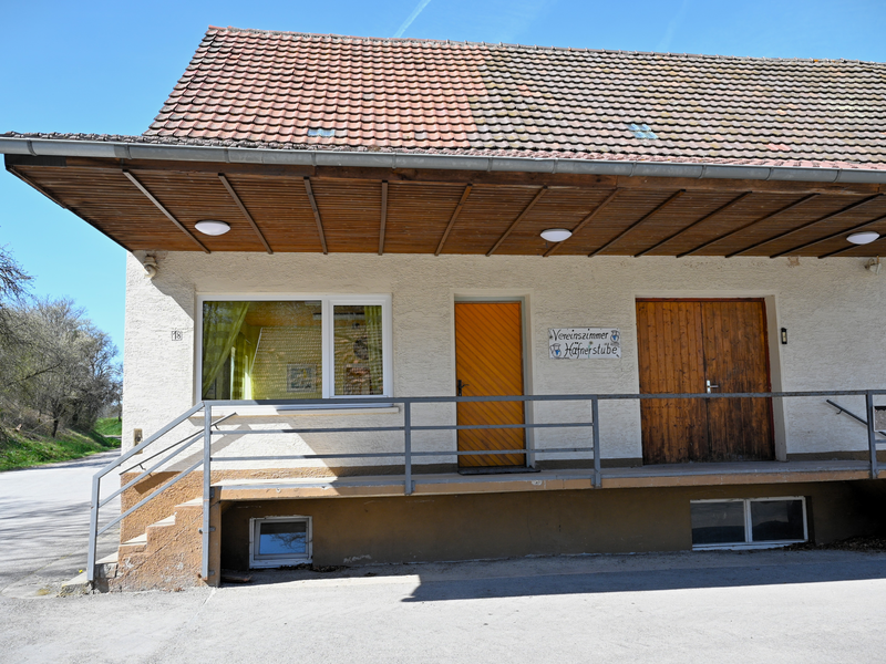 Ein Gebäude mit einem schrägen Ziegeldach, einer Rampe und einer Holztür, auf der ein Schild mit der Aufschrift „Vereinszimmer Häfnerstube“ angebracht ist.