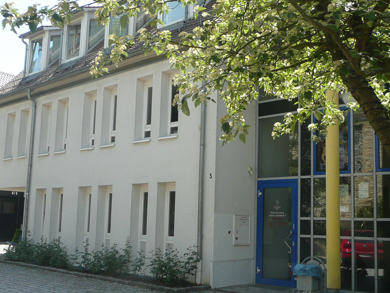 Ein zweigeschossiges Gebäude mit weißen Wänden und großen Fenstern, umgeben von einem gepflasterten Hof und einem Baum im Vordergrund.