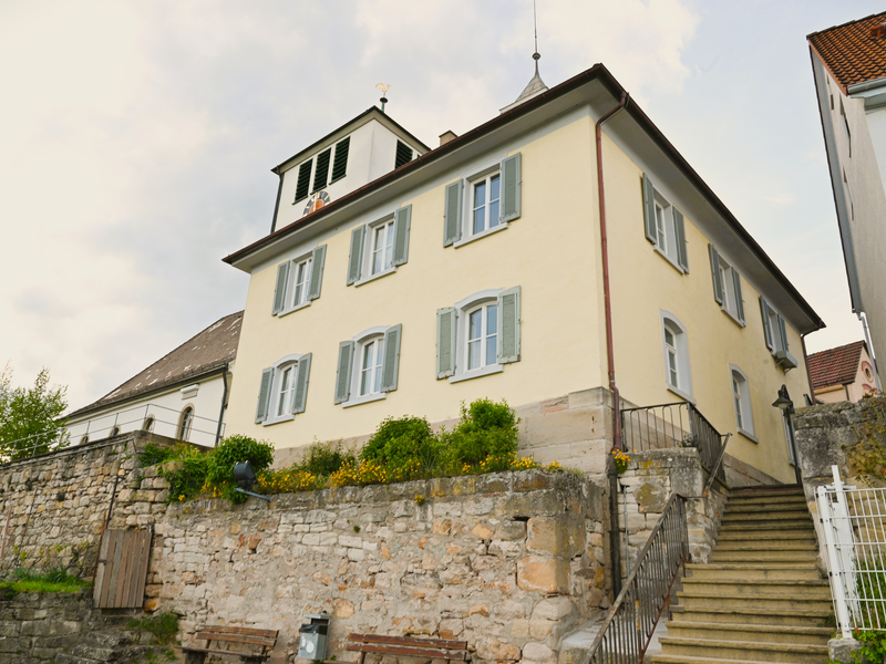 Ein zweistöckiges, hellgelbes Haus mit grünen Fensterläden steht auf einem erhöhten Fundament neben einer Steintreppe und einer Steinmauer.