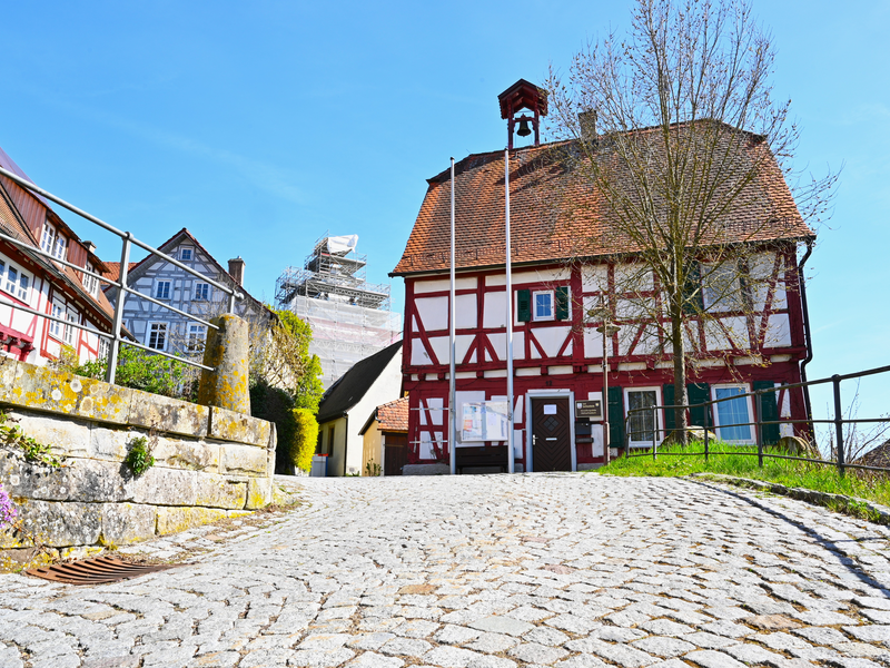 Das Bild zeigt ein malerisches Fachwerkhaus mit roten Balken und weißer Fassade auf einem gepflasterten Weg, umgeben von mehreren weiteren Gebäuden und einem blauen Himmel im Hintergrund.
