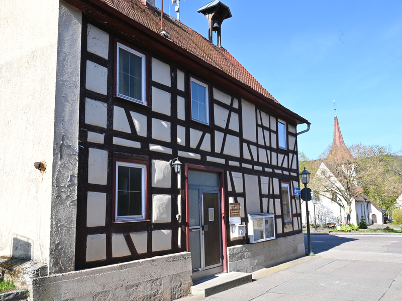 Ein traditionelles Fachwerkhaus mit braunen Balken und weißen Wänden steht in einer sonnigen Straßenszene, im Hintergrund ist eine Kirche sichtbar.
