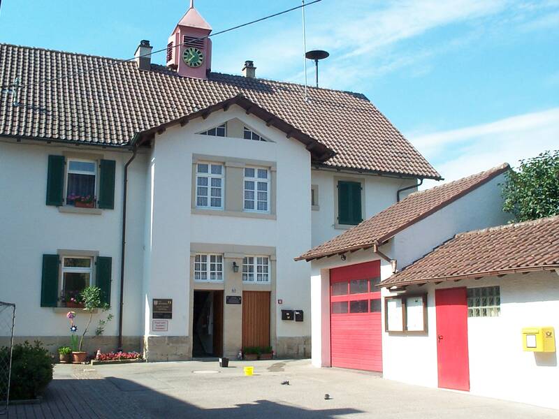 Ein zweistöckiges Gebäude mit rotem Ziegeldach, einer Uhr im Dachgiebel und angrenzenden Garagen mit roten Toren.