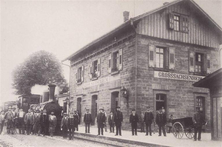 Vor einem historischen Bahnhofsgebäude mit der Aufschrift "Großsachsenheim" stehen mehrere uniformierte Personen und weitere Männer neben einer alten Dampflokomotive.