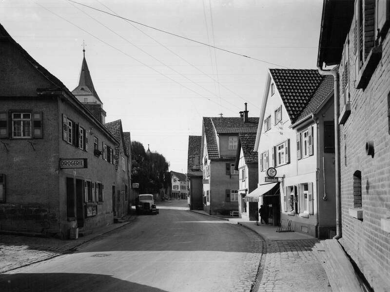 Eine historische Straßenansicht zeigt eine ruhige deutsche Kleinstadt mit Fachwerkhäusern, einem Drogeriegeschäft links und einer Kirche im Hintergrund.
