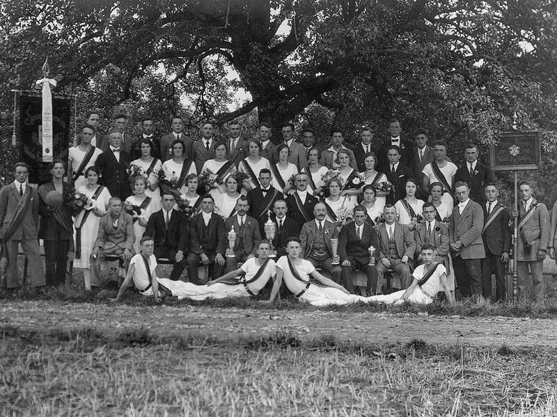 Eine große Gruppe von Männern und Frauen, teils in formeller Kleidung und teils in Sportbekleidung, posiert im Freien vor einem Baum.