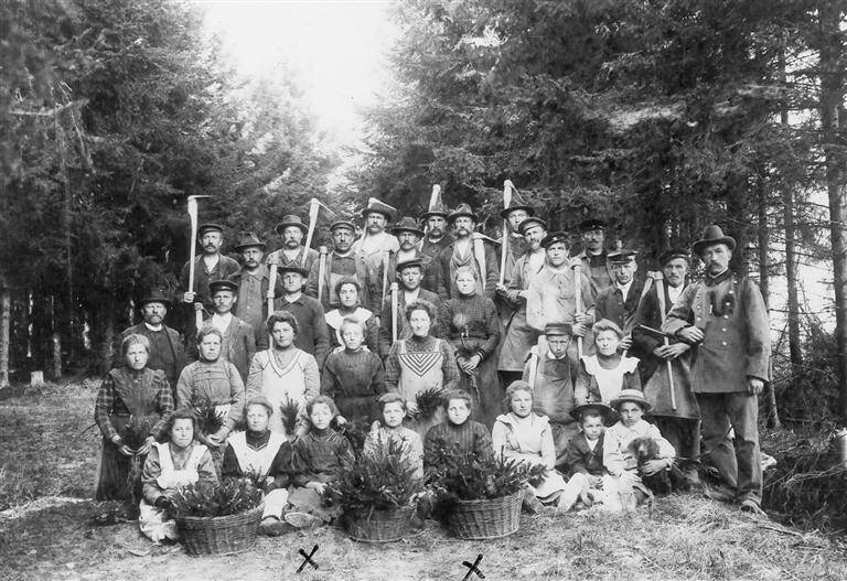 Eine Gruppe von Männern, Frauen und Kindern posiert in traditioneller Kleidung mit Werkzeugen und Körben voller Zweige im Wald.